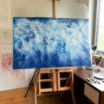 Ocean Blues watercolor painting on an easel in the artist's studio. The abstract seascape features tranquil blue tones and foam details, showcasing the creative process of Kateryna Nazarenko.