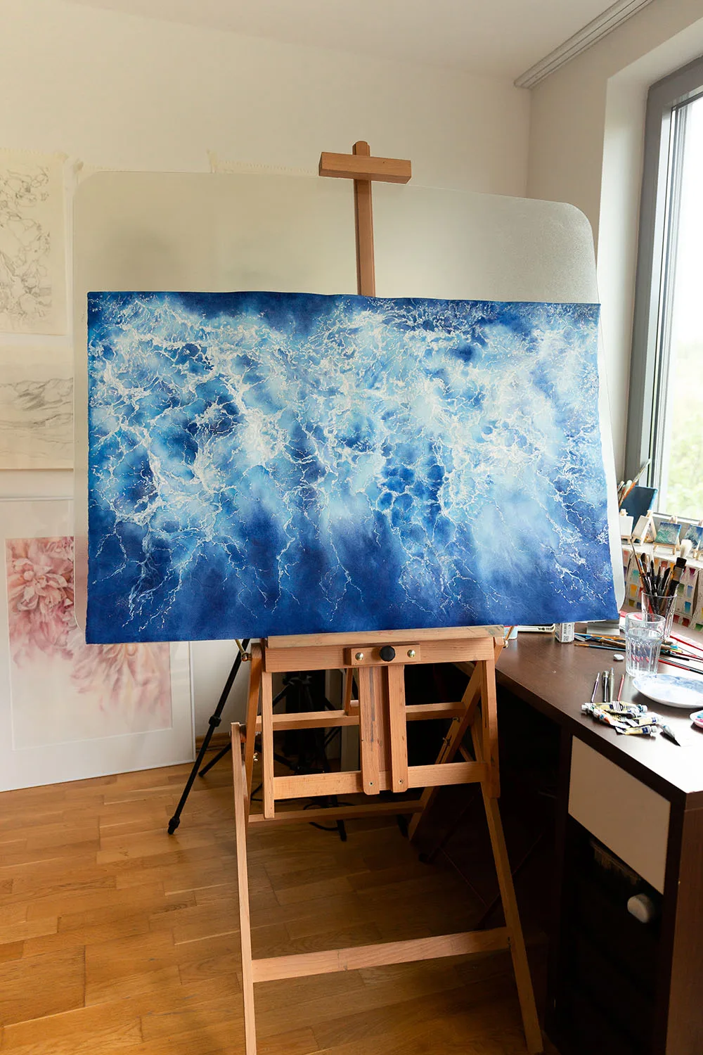 Ocean Blues watercolor painting on an easel in the artist's studio. The abstract seascape features tranquil blue tones and foam details, showcasing the creative process of Kateryna Nazarenko.