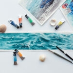 ‘Sparkles on Turquoise Water’ in progress on a table in Kateryna Nazarenko’s art studio, surrounded by watercolor brushes and palettes, showcasing the delicate layers of turquoise and shimmering light reflections taking shape.