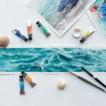 ‘Sparkles on Turquoise Water’ in progress on a table in Kateryna Nazarenko’s art studio, surrounded by watercolor brushes and palettes, showcasing the delicate layers of turquoise and shimmering light reflections taking shape.