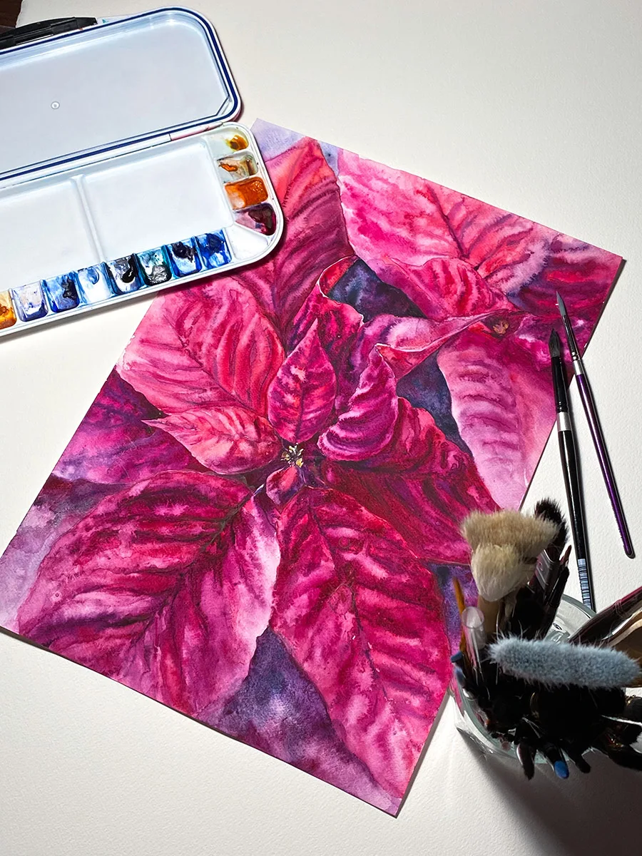 Magenta Poinsettia watercolor painting displayed on a desk surrounded by paints and brushes, reflecting the creative atmosphere of an artist's workspace, perfect for adding vibrant, festive energy to your studio.