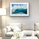 Framed 'Through the Light' watercolor painting above a sofa in a coastal living room, capturing the motion of a powerful ocean wave in deep blues and turquoise, enhancing the serene yet dynamic coastal ambiance of the space.