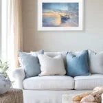 Framed fine art print of Morning Solitude watercolor painting with boat and ocean sunrise, displayed above sofa in a beach-style coastal living room interior.