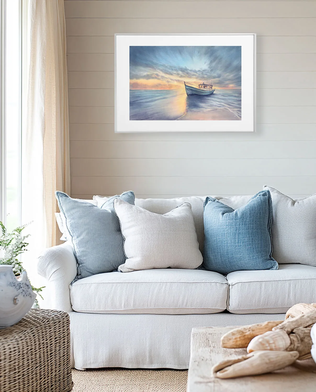 Framed fine art print of Morning Solitude watercolor painting with boat and ocean sunrise, displayed above sofa in a beach-style coastal living room interior.